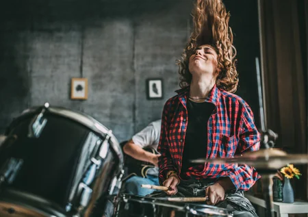 A young person sitting at a drum set in what appears to be a recording studio or rehearsal space. They have long, curly hair and are wearing a plaid flannel shirt. The person has a focused, thoughtful expression on their face as they sit behind the drums, suggesting they are engaged in the act of drumming or musical performance.